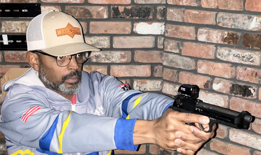 A man wearing glasses and a baseball cap is holding a handgun equipped with a red dot sight.