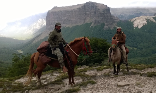 Two men on horseback are pictured in a mountainous, rugged landscape. They are dressed for outdoor exploration or hunting, with gear packed on their horses.