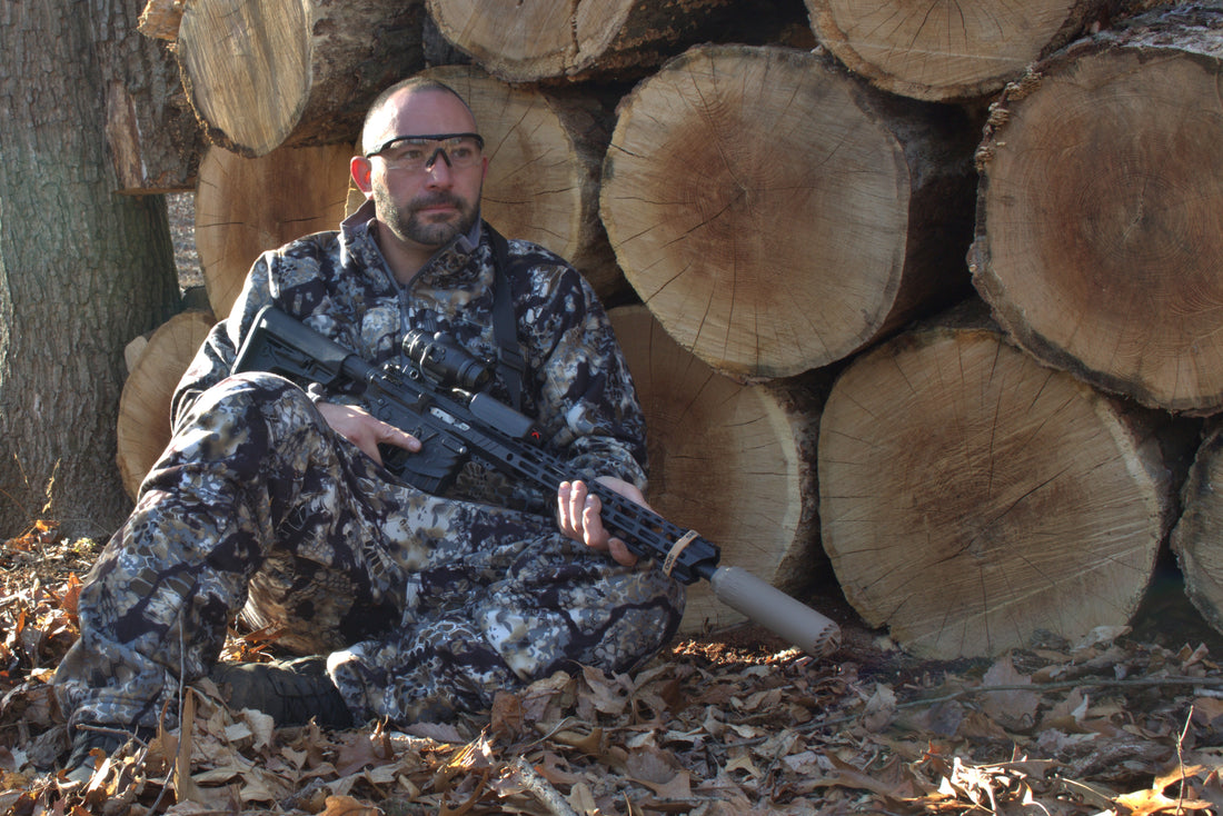 A hunter dressed in camouflage gear sits on the ground, leaning against a stack of large, cut logs. He is holding a rifle equipped with a suppressor, scope, and other tactical accessories. 