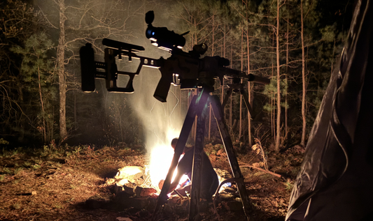 A tactical rifle equipped with a thermal scope is mounted on a tripod backlit by a campfire.