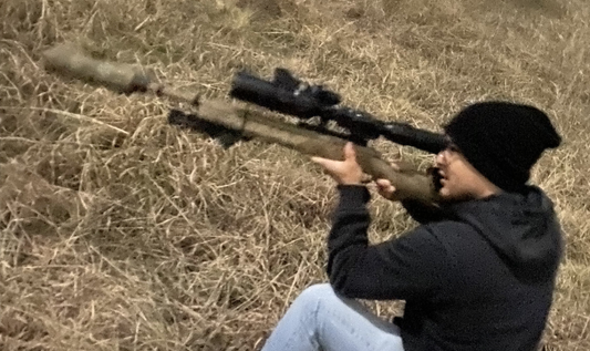 A person wearing a black beanie and dark clothing is aiming a long-range rifle with a suppressor and a large scope while sitting in a grassy field. 