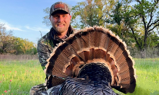 A hunter dressed in camouflage proudly displays a large turkey, showcasing the bird's impressive fan of tail feathers. 