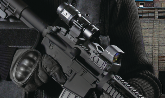 The image shows a close-up of a tactical rifle equipped with multiple optics, including a red dot sight and a magnifier. 