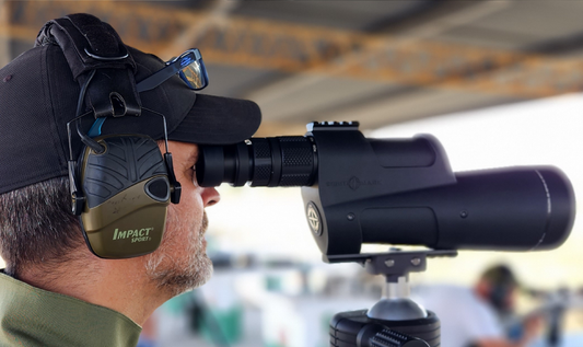 A man is using a spotting scope at a shooting range while wearing ear protection and sunglasses.